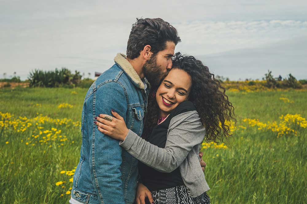 Man kissing a smiling woman's forehead in a field of yellow flowers.