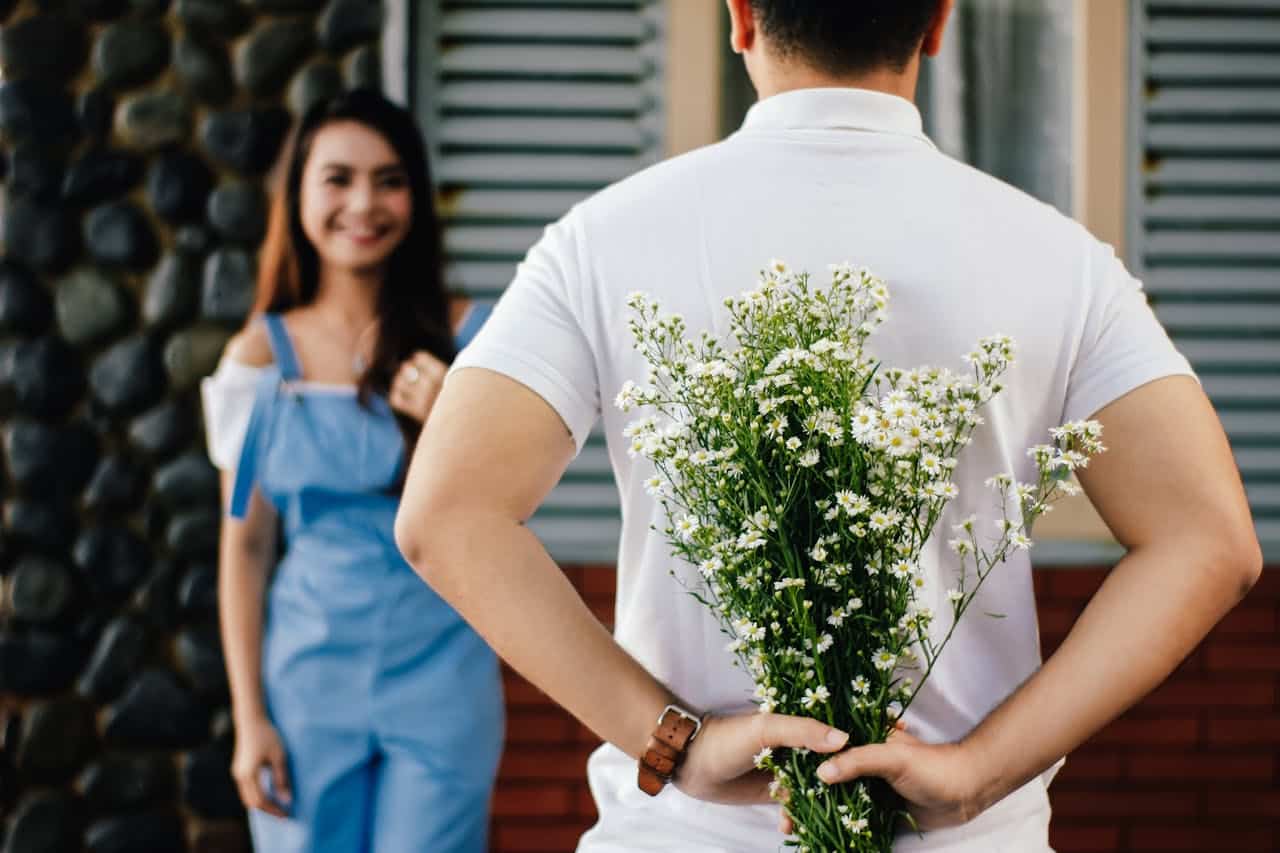 Man holding flowers behind his back while smiling woman waits in the background