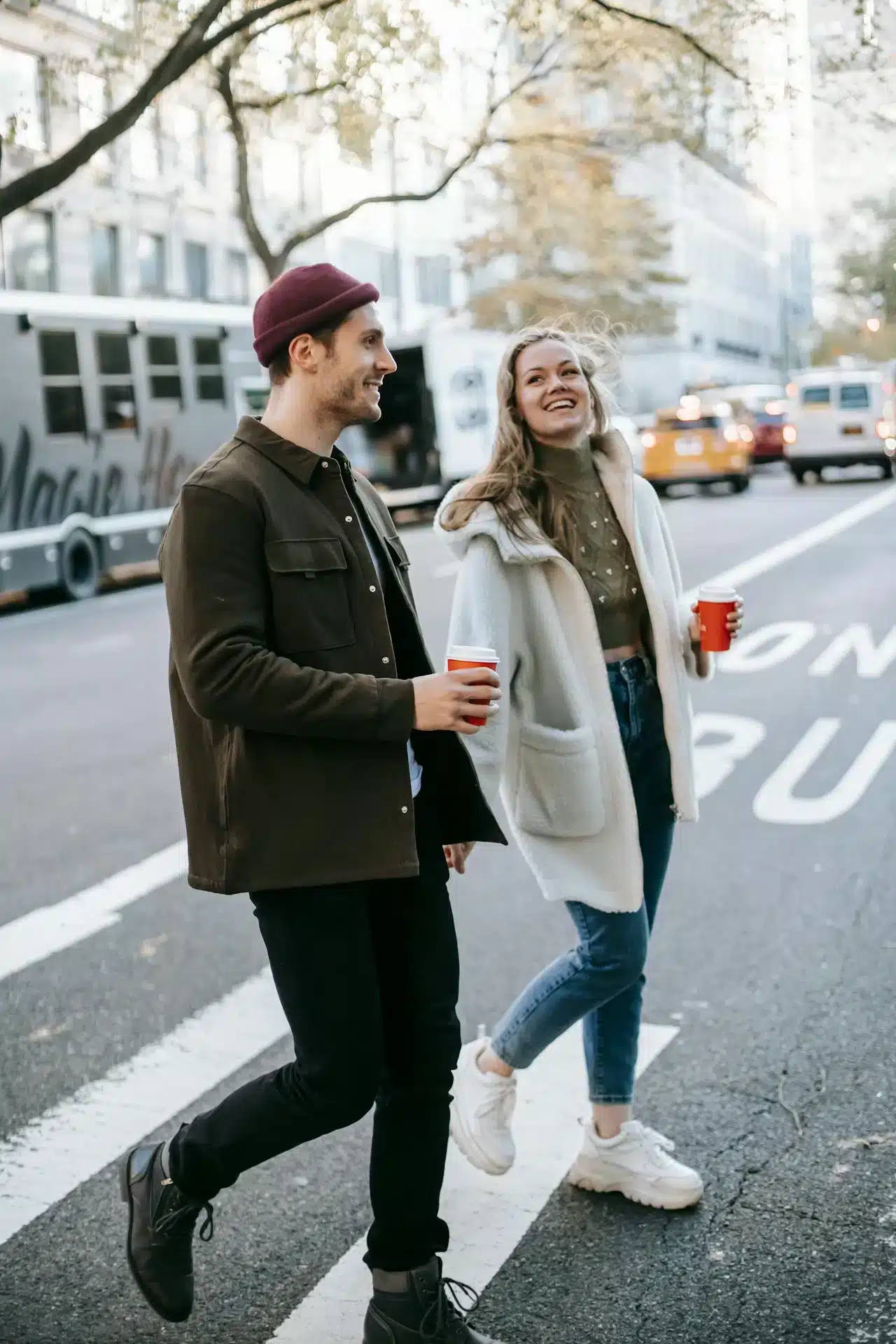 A couple crosses a Massachusetts street holding coffee cups, smiling at each other while walking in an urban setting.
