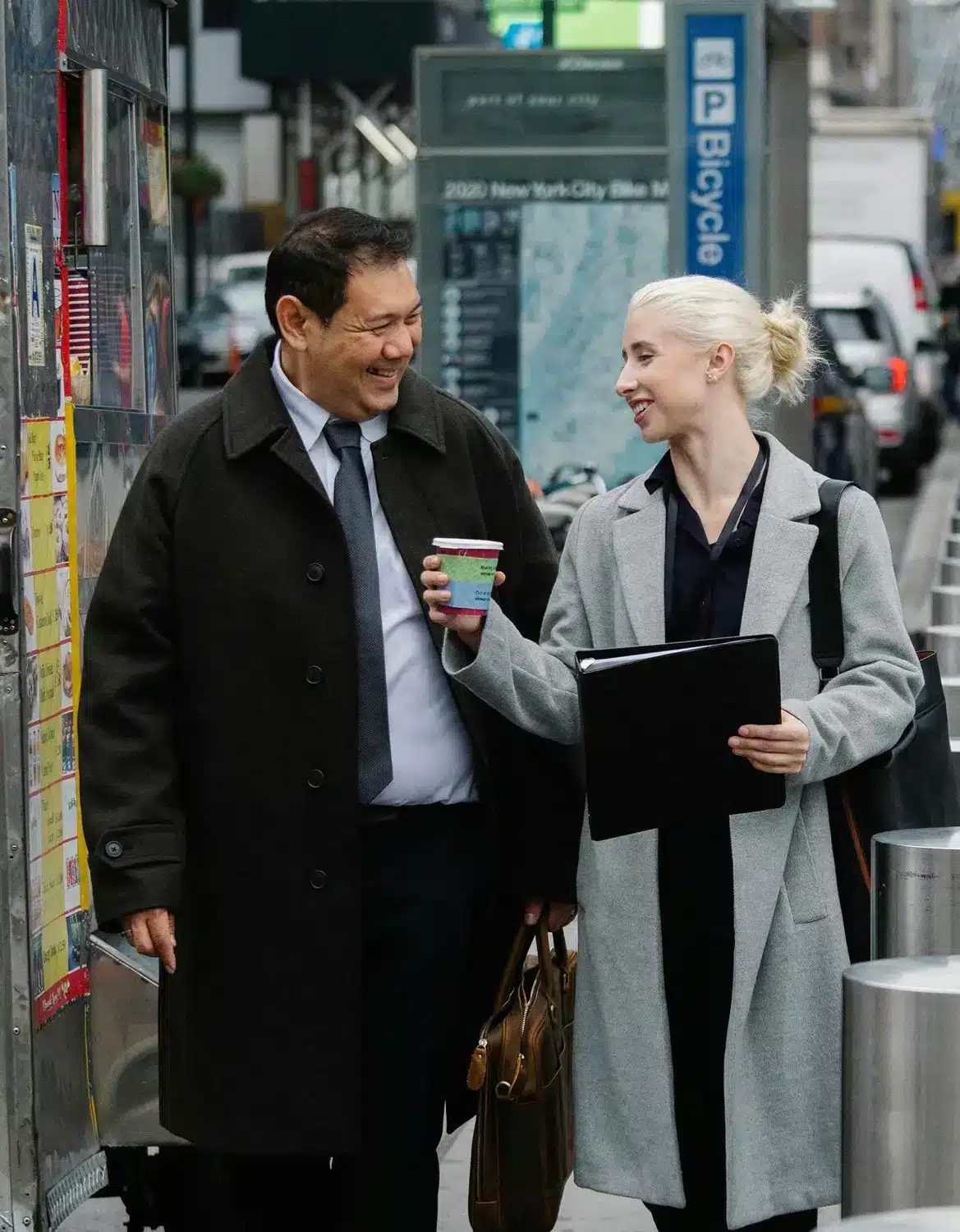 Asian man and a white woman walk side by side on a city street, smiling warmly as she carries coffee and a work folder.