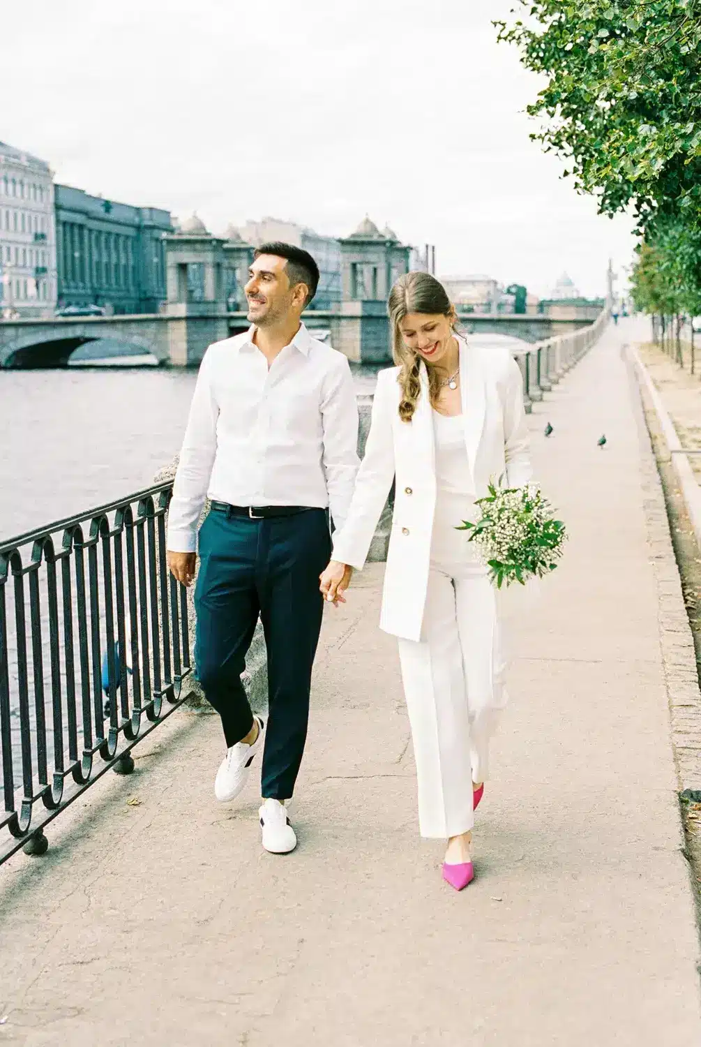 A couple walks across a bridge beside water and city buildings, smiling as the woman holds flowers in her hand.