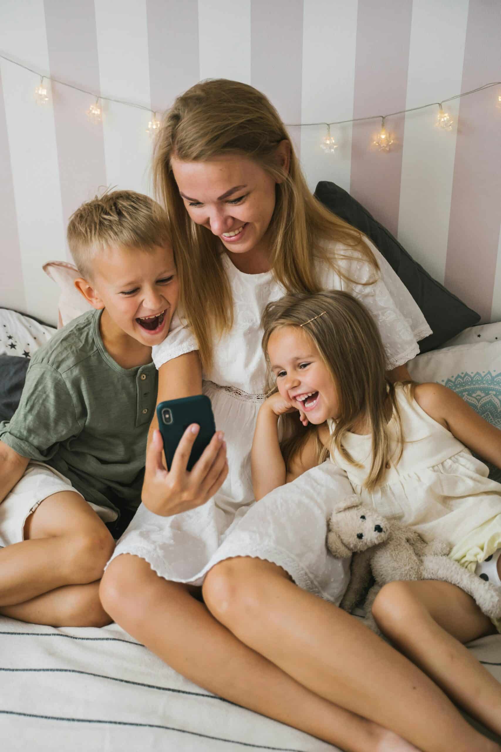Mother sitting with two children as they watch a phone together, smiling and sharing a playful moment