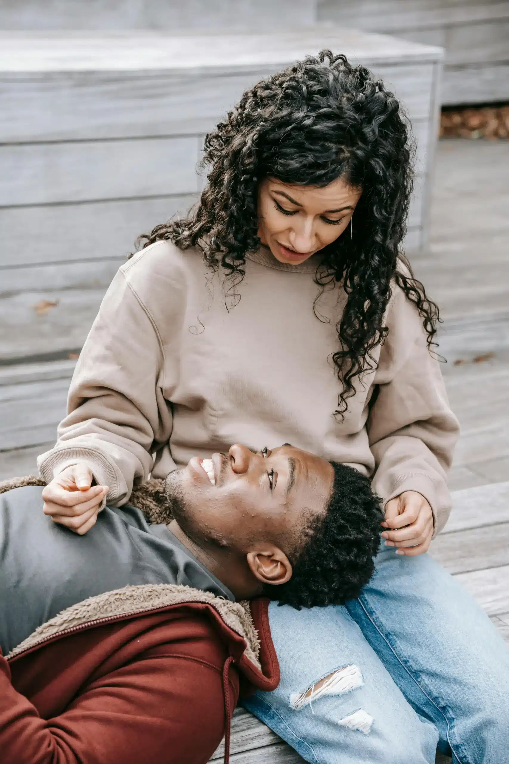  A couple relaxing on a wooden patio, the man resting his head in his partner’s lap as they talk and connect calmly