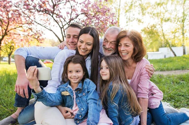 Three-generation family sitting outside in nature taking a selfie with a smartphone.