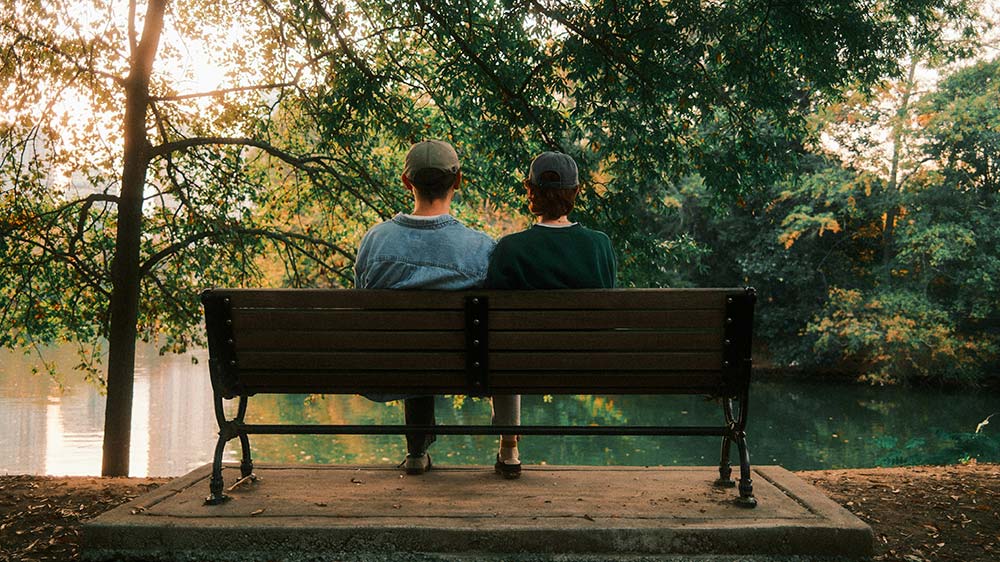 Two people sitting on a bench looking at the water in a calm outdoor setting.