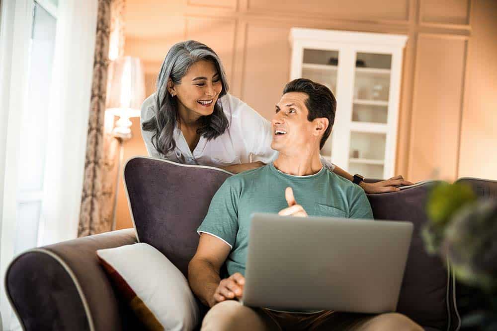 Waist-up of a happy couple in love using a laptop while sitting in a living room.