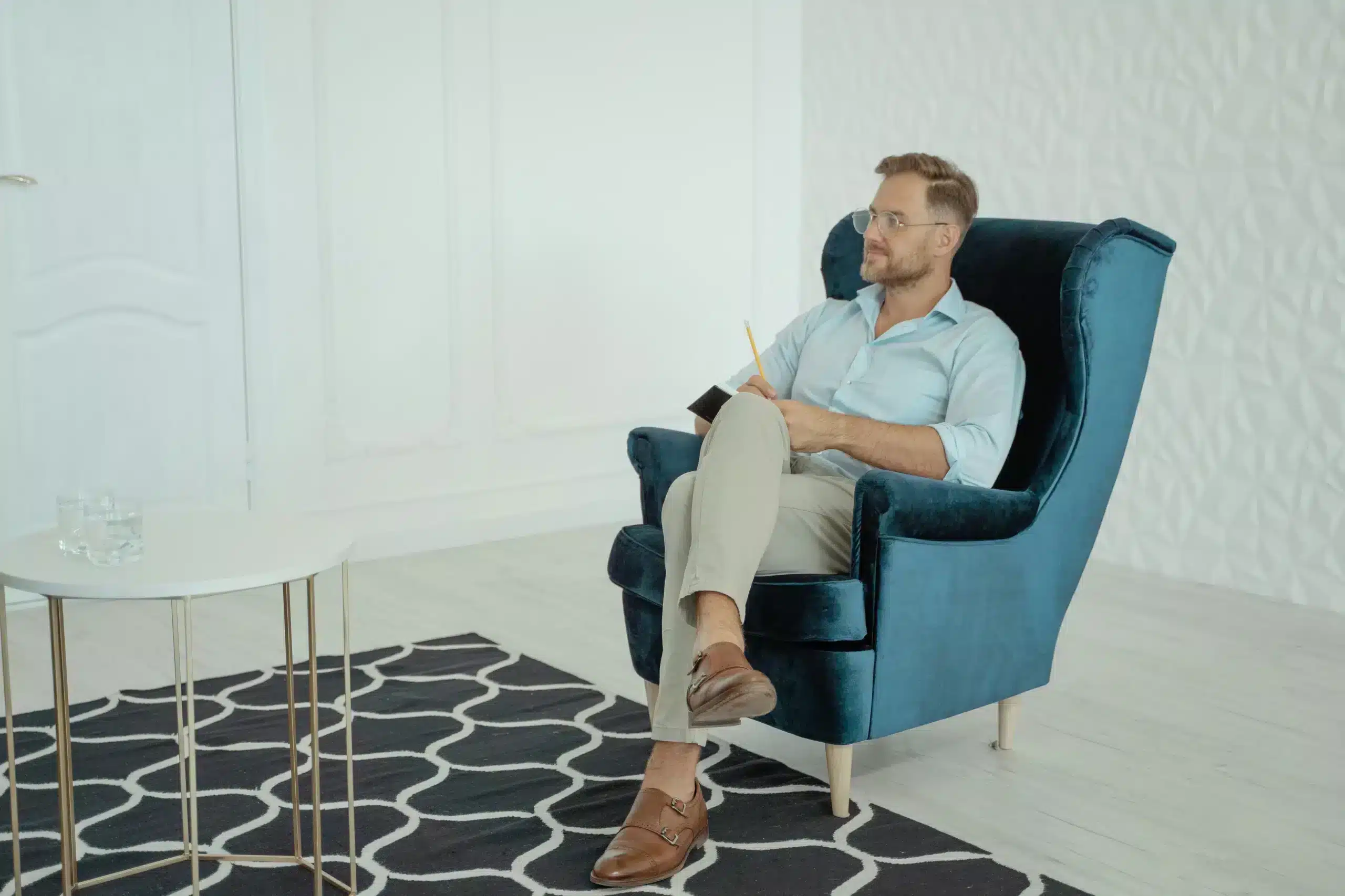 A professional man with glasses sits cross-legged in a blue wingback armchair, holding a notebook and pencil while listening attentively in an office.