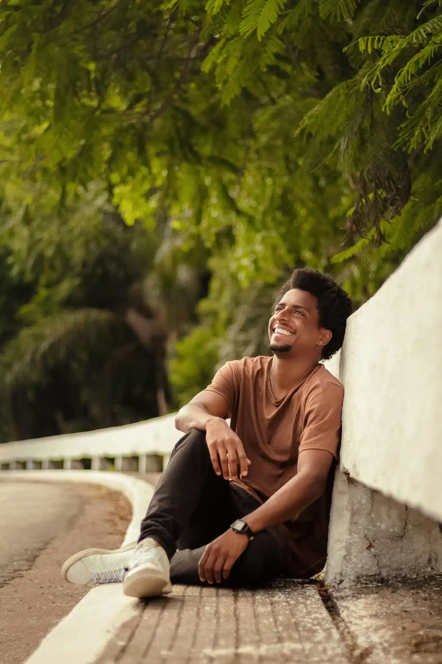 A smiling young Black man sits on a roadside curb, leaning against a low white wall while looking up thoughtfully under a canopy of green trees.