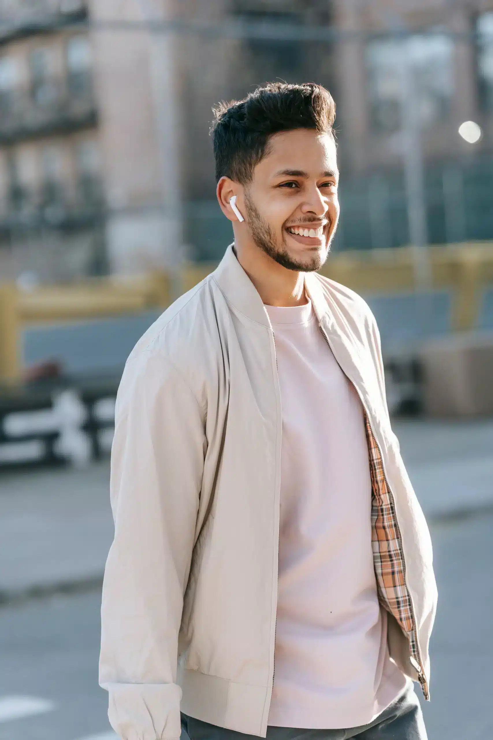 A smiling young man with a beard and wireless earbuds walks outdoors, wearing a light-colored bomber jacket over a pink t-shirt.