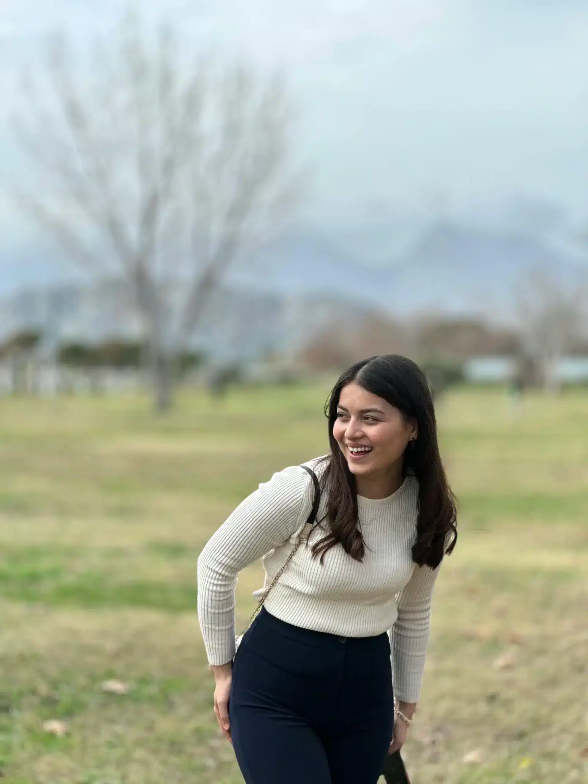 A young woman with long dark hair smiles and looks aside in a grassy field, wearing a cream ribbed sweater and dark trousers with a blurred tree in the background.