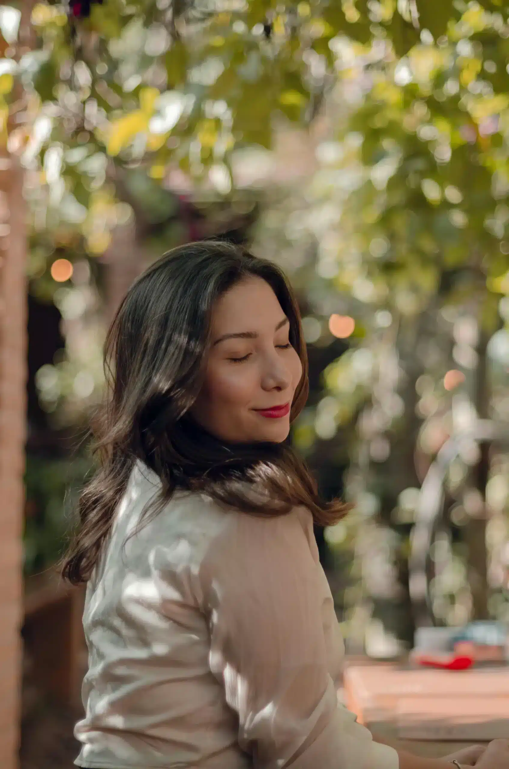 A young woman with dark hair and red lipstick smiles peacefully with her eyes closed against a blurred, sunlit green background.