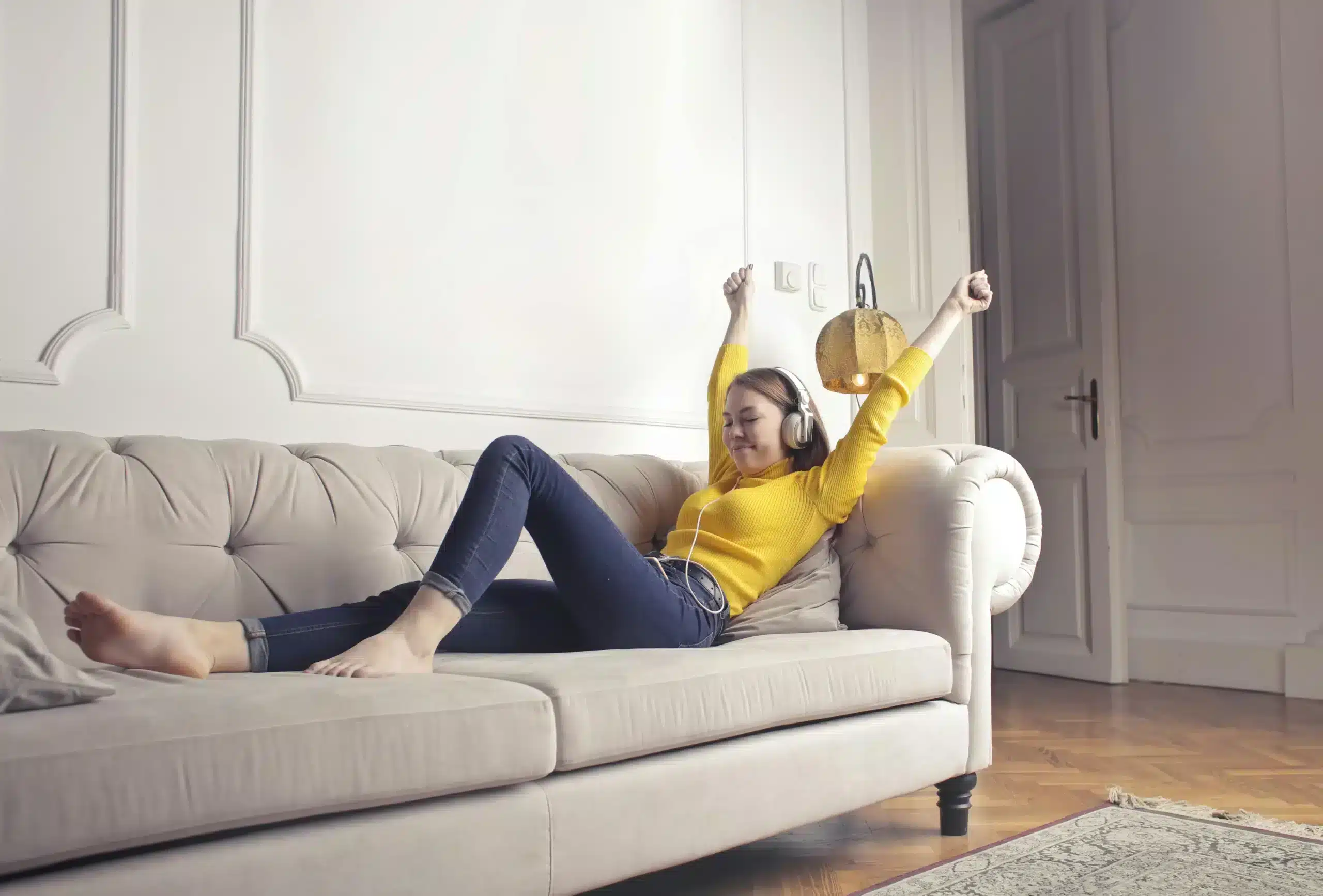 A young woman in a yellow sweater and jeans relaxes on a tufted sofa, stretching her arms up while listening to music with headphones.