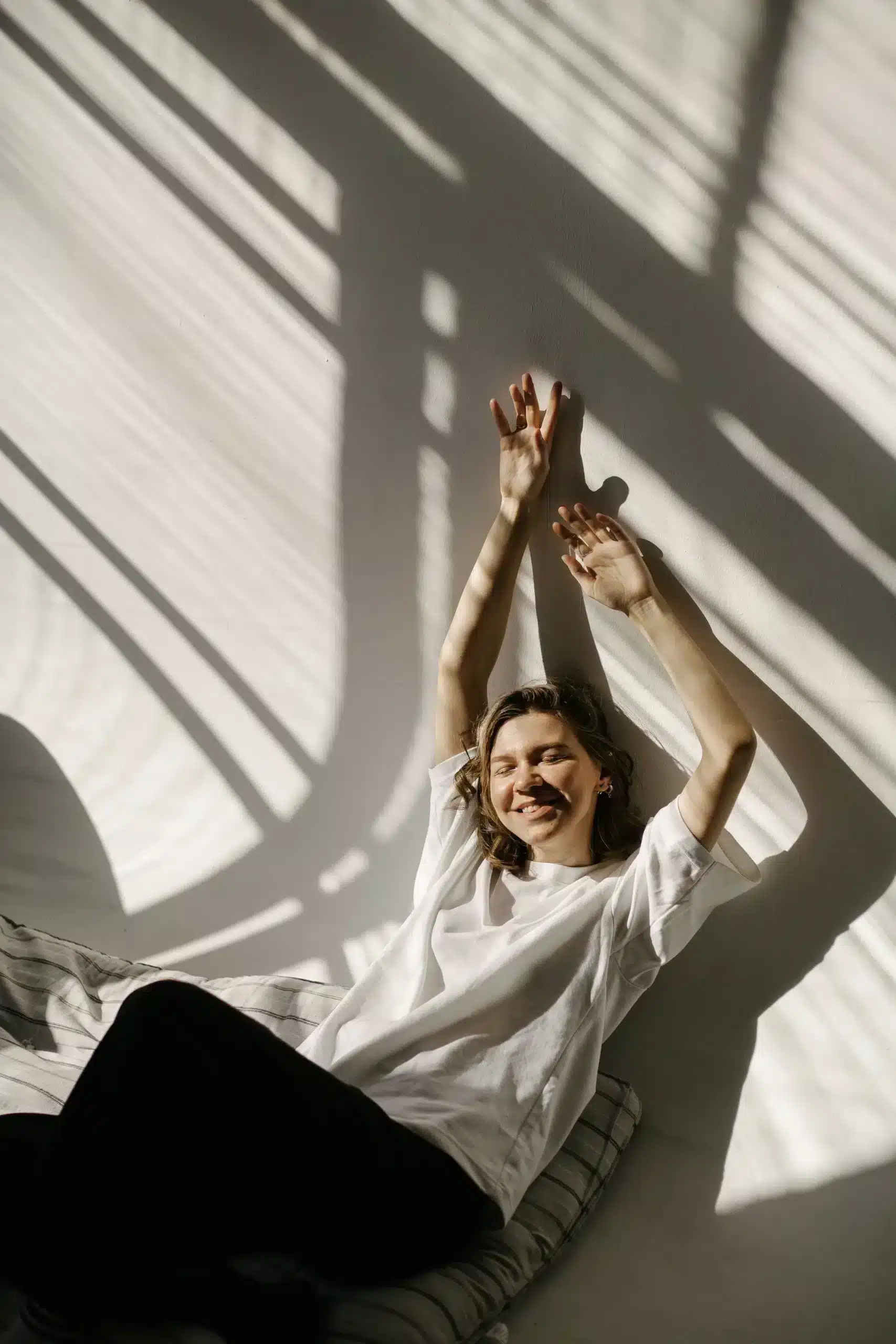 A smiling young woman sits on a floor cushion, stretching her arms upward against a white wall illuminated by dramatic, striped window shadows