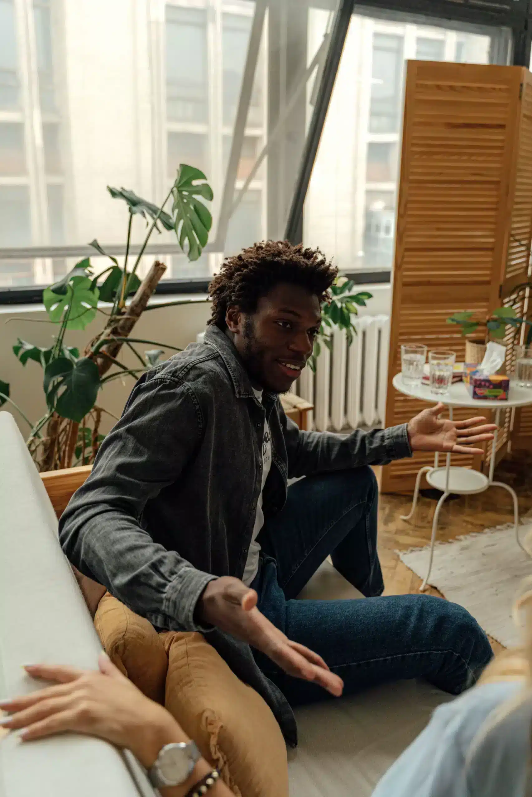 A young Black man with curly hair sits on a couch, gesturing with his hands while engaged in an animated conversation in a bright, plant-filled room.